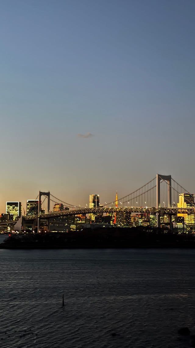 Un grande ponte sospeso illuminato attraversa l'acqua con uno skyline cittadino e una torre rossa al tramonto.
