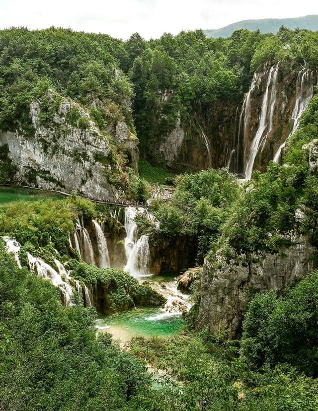 Une série de cascades dévale des formations rocheuses moussues dans un bassin turquoise, le tout entouré d'une dense forêt verte et d'une passerelle où se trouvent des promeneurs.