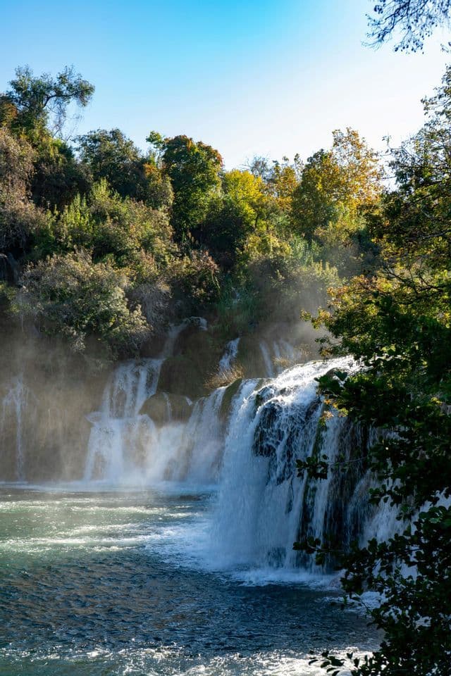 Une vaste cascade se déverse dans une rivière, créant une brume sous le soleil. Elle est entourée d'une forêt dense sous un ciel bleu pur.