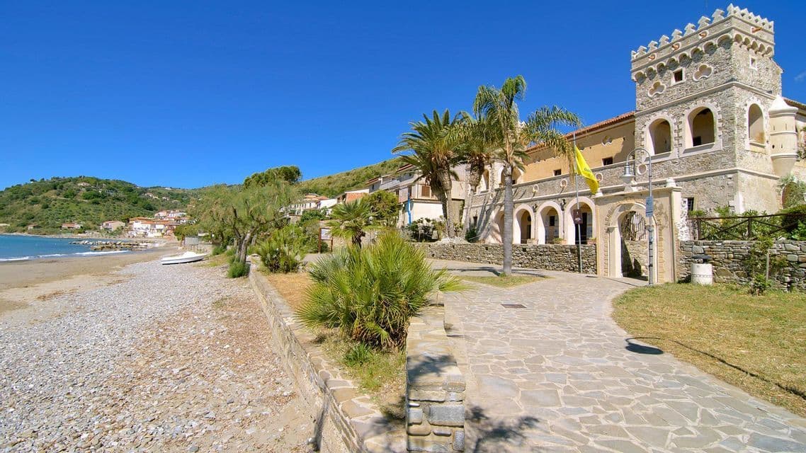 Un sentiero lastricato in pietra costeggia una spiaggia di ciottoli, dirigendosi verso un edificio costiero simile a un castello, sotto un cielo azzurro e limpido.