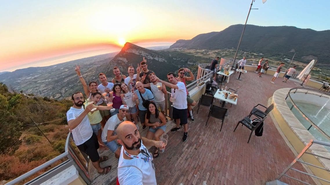 Un viaggio di gruppo WeRoad scatta un selfie su una terrazza panoramica, brindando con drink al tramonto in montagna.