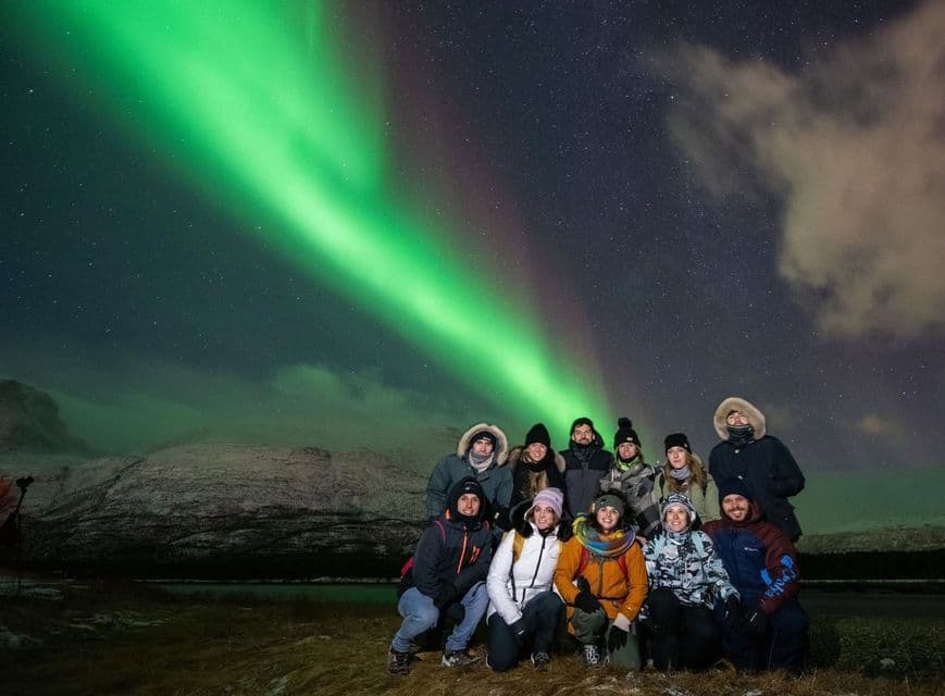 Un grupo de viaje de WeRoad posa junto por la noche, con la aurora boreal verde iluminando el cielo estrellado sobre ellos.