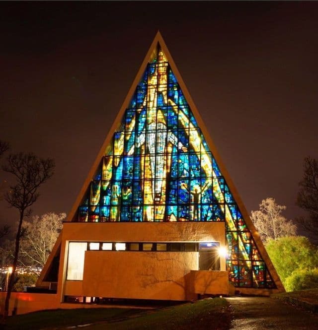 Un moderno edificio con forma de A, iluminado por la noche, que presenta un enorme y colorido vitral que forma su fachada.