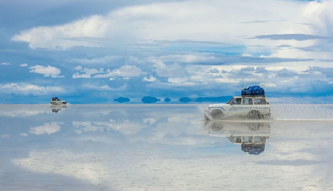 Two white off-road vehicles drive through a vast, shallow body of water that perfectly reflects the cloudy sky.