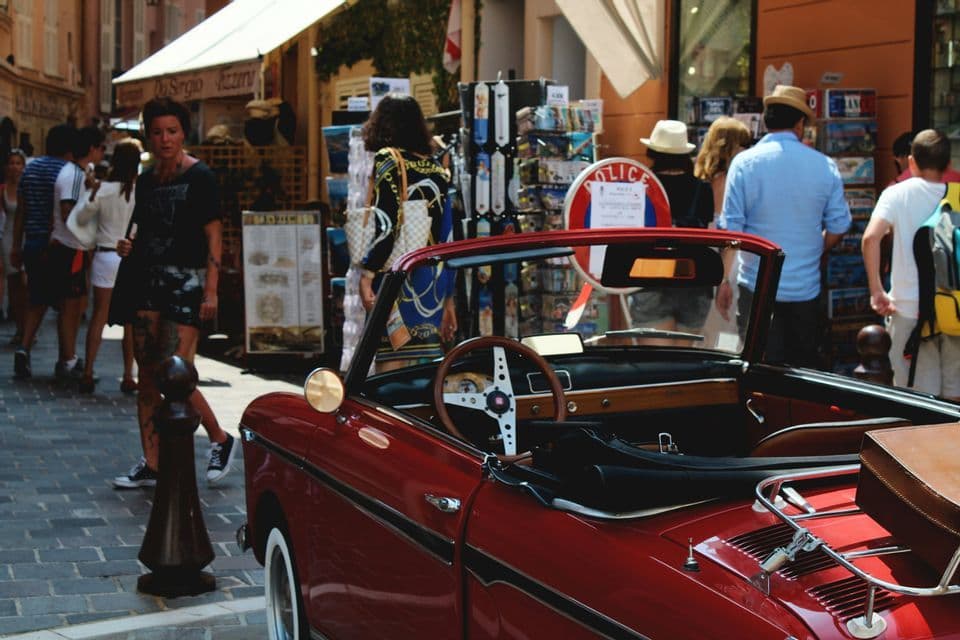 Una classica auto cabrio rossa è parcheggiata su una trafficata strada di ciottoli con persone che passeggiano davanti ai negozi sullo sfondo.