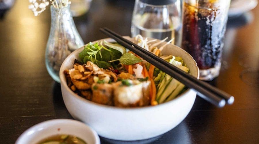 Nahaufnahme einer weißen Schale mit asiatischem Nudelsalat und Essstäbchen, auf einem dunklen Tisch mit einem Glas Limo im Hintergrund.