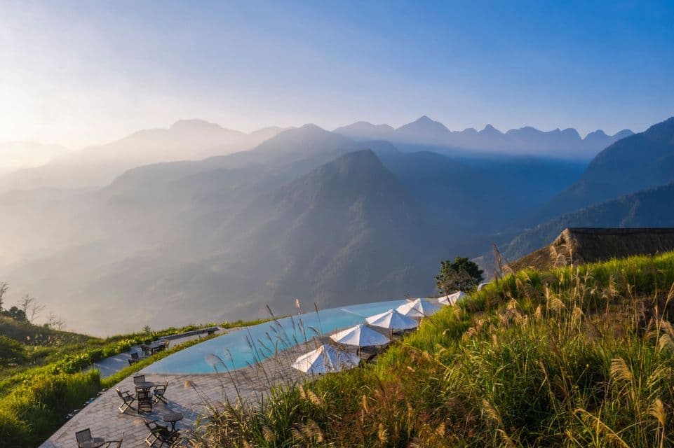 Ein Infinity-Pool auf einem grasbewachsenen Hügel mit Tischen und Sonnenschirmen blickt bei Sonnenaufgang auf eine weite, dunstige Bergkette.