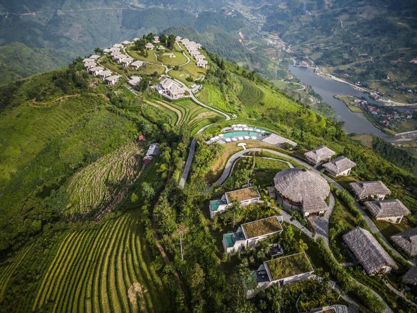 Vista aérea de un resort con villas y piscinas en una exuberante montaña verde, con vistas a campos en terrazas y un valle fluvial.