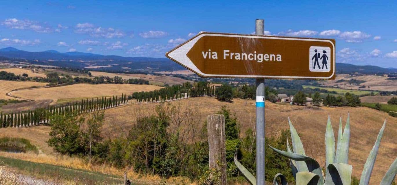 Un cartello marrone della Via Francigena indica una valle di dolci colline, campi e cipressi sotto un cielo azzurro.