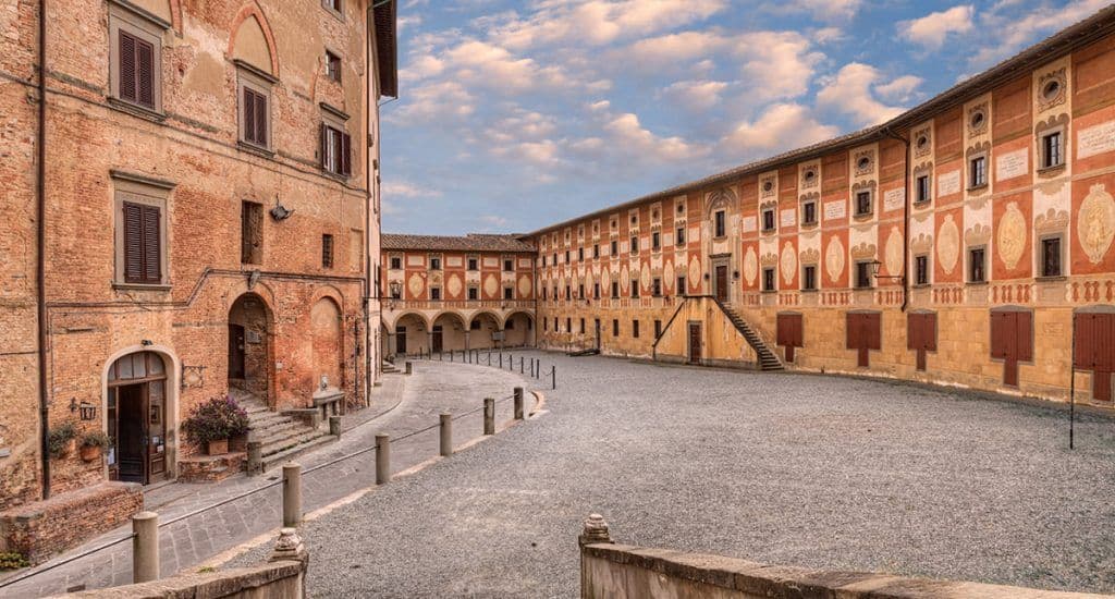 Un grande cortile vuoto con superficie in ghiaia è circondato da edifici storici in mattoni e dipinti sotto un cielo blu nuvoloso.