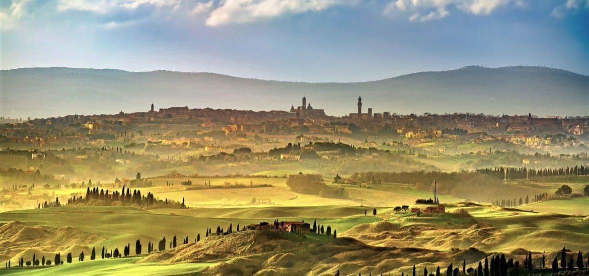 Una vista panoramica di una città storica su una collina, osservata attraverso un paesaggio nebbioso di dolci campi verdi e cipressi.
