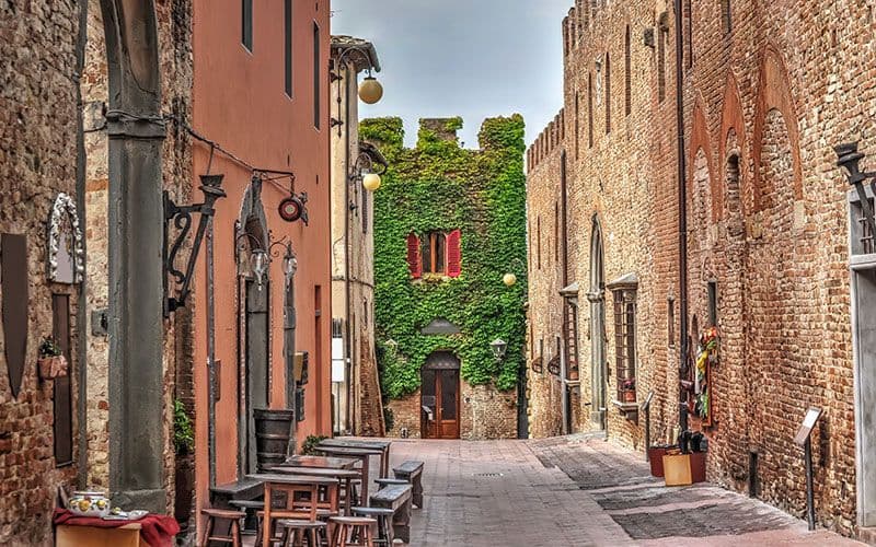 Un vicolo medievale stretto e vuoto tra edifici in mattoni, con una torre ricoperta d'edera e persiane rosse in fondo.