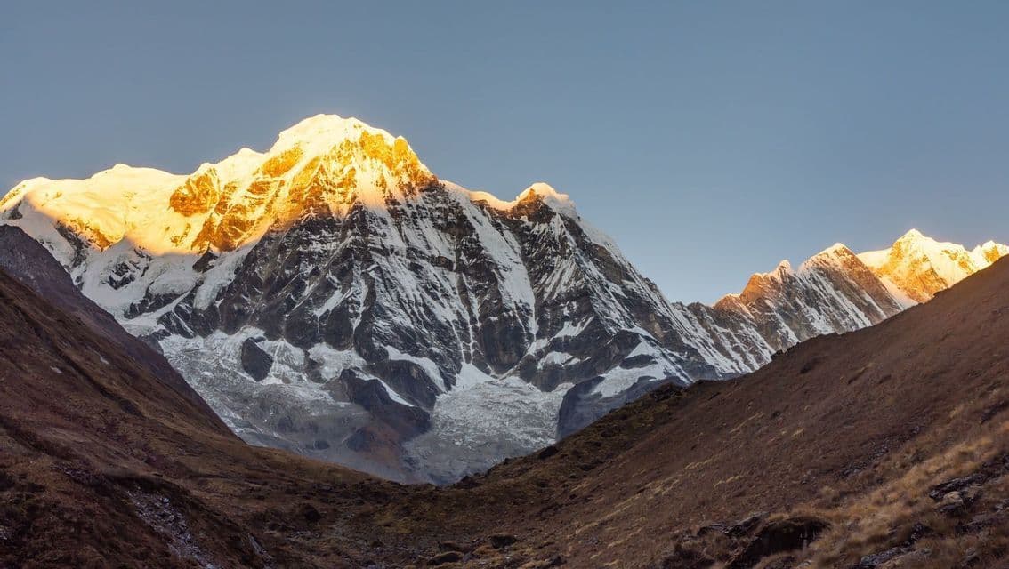 Le sommet d'une grande montagne enneigée est illuminé par la lumière dorée du soleil contre un ciel bleu clair, vu depuis une vallée rocheuse.