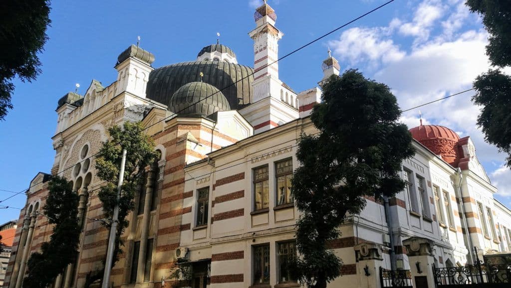 Una sinagoga riccamente decorata con diverse cupole scure, mattoni a strisce e una cupola rossa, ripresa dal basso contro un cielo blu.