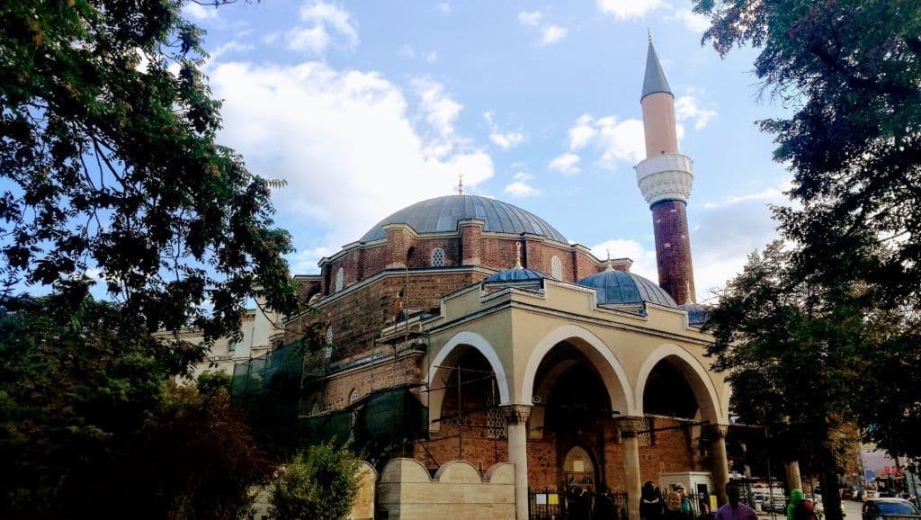 Una grande moschea in mattoni con una cupola principale e un alto minareto, incorniciata da alberi verdi sotto un cielo blu con nuvole bianche.