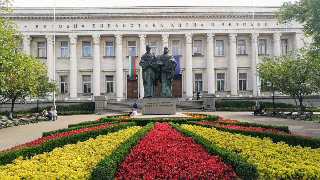 Aiuole colorate in un giardino formale conducono a una grande statua di bronzo di fronte a un edificio neoclassico con colonne.