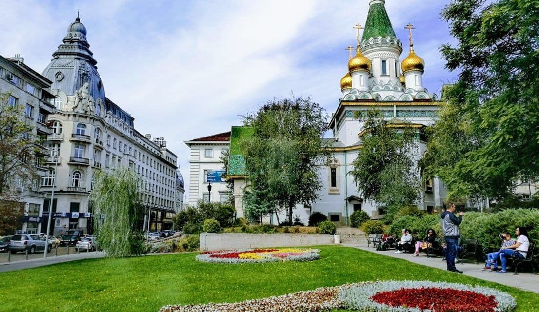 Un parco cittadino con aiuole colorate, con una chiesa ornata dalle cupole dorate e un edificio storico sullo sfondo.