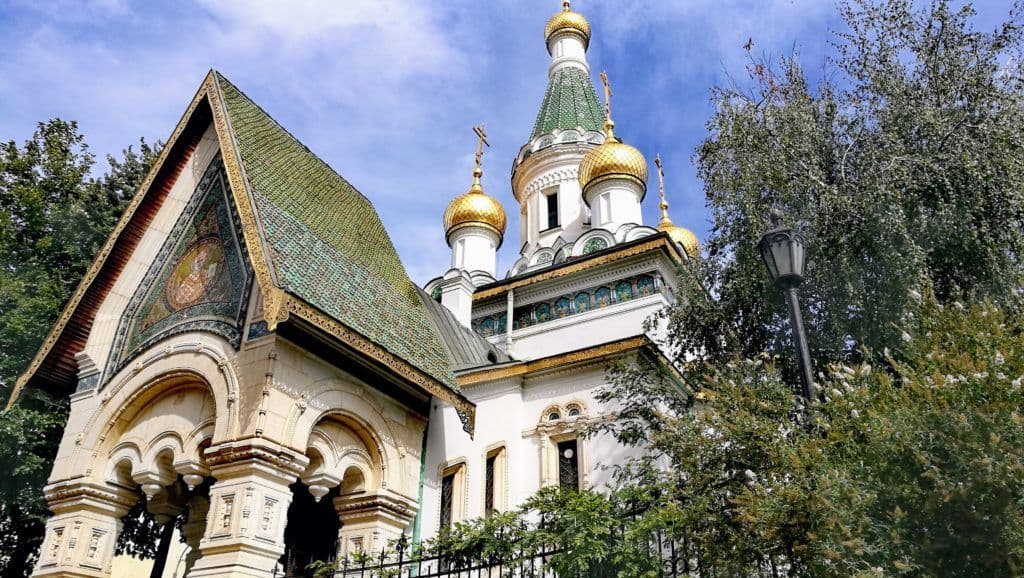 Una chiesa ortodossa bianca con cupole a cipolla dorate, un tetto di tegole verdi e un mosaico si erge dietro gli alberi sotto un cielo azzurro.