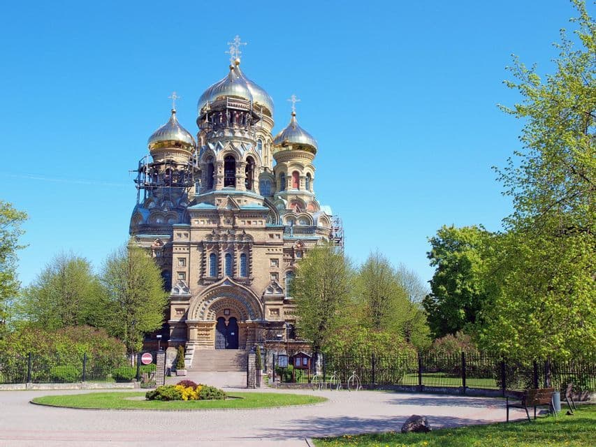 Una sfarzosa cattedrale ortodossa con cupole dorate e impalcature si erge dietro un parco con alberi verdi sotto un cielo limpido.