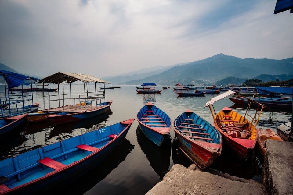 Colorful boats moored on a calm lake with hazy mountains in the background under a cloudy sky.