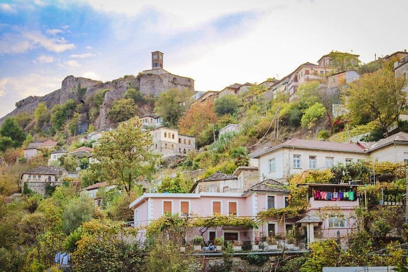 Un pueblo en la ladera con casas tradicionales y exuberante vegetación que asciende hasta una fortaleza de piedra con torre del reloj.