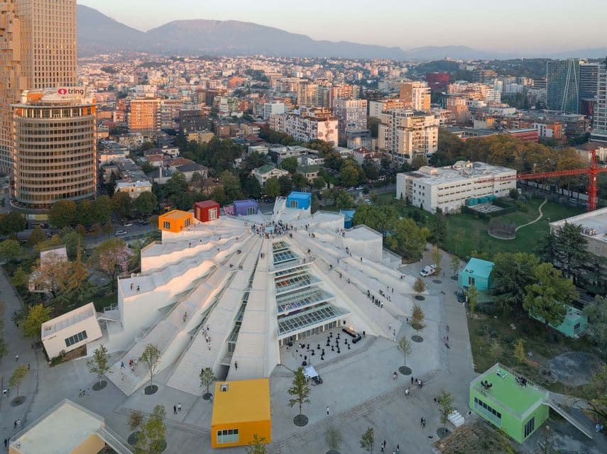 Vista aérea de un edificio moderno, blanco, con forma de pirámide, con estructuras coloridas en forma de caja en la parte superior, en un paisaje urbano al atardecer.