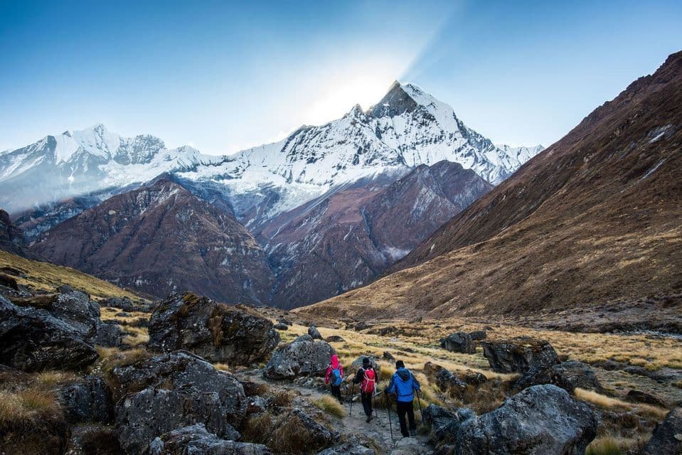 Un voyage de groupe WeRoad en randonnée sur un sentier rocailleux vers une montagne massive enneigée, alors que le soleil se lève derrière son sommet.