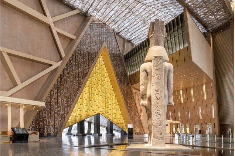 Una colosal estatua del antiguo Egipto se alza en una sala de museo moderna junto a una pared triangular decorada con oro y jeroglíficos.