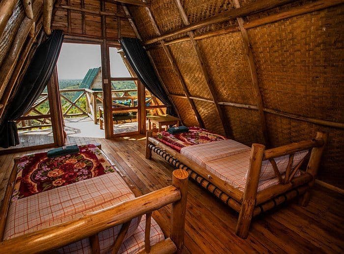 L'interno di una rustica baita a capanna con due letti in legno, pareti intrecciate e una porta aperta che si affaccia su un balcone e colline verdi.