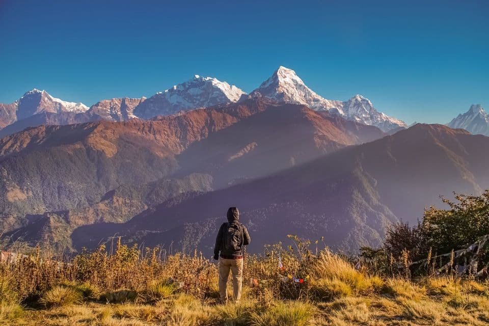 Una persona con uno zaino si trova su una collina erbosa, ammirando una vasta catena montuosa con cime innevate sotto un cielo azzurro e limpido.