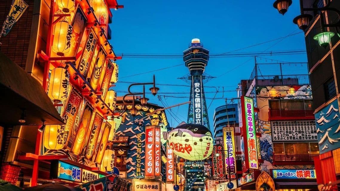 Una calle japonesa al atardecer con farolillos luminosos, un cartel de pez globo y una torre iluminada contra un cielo azul.
