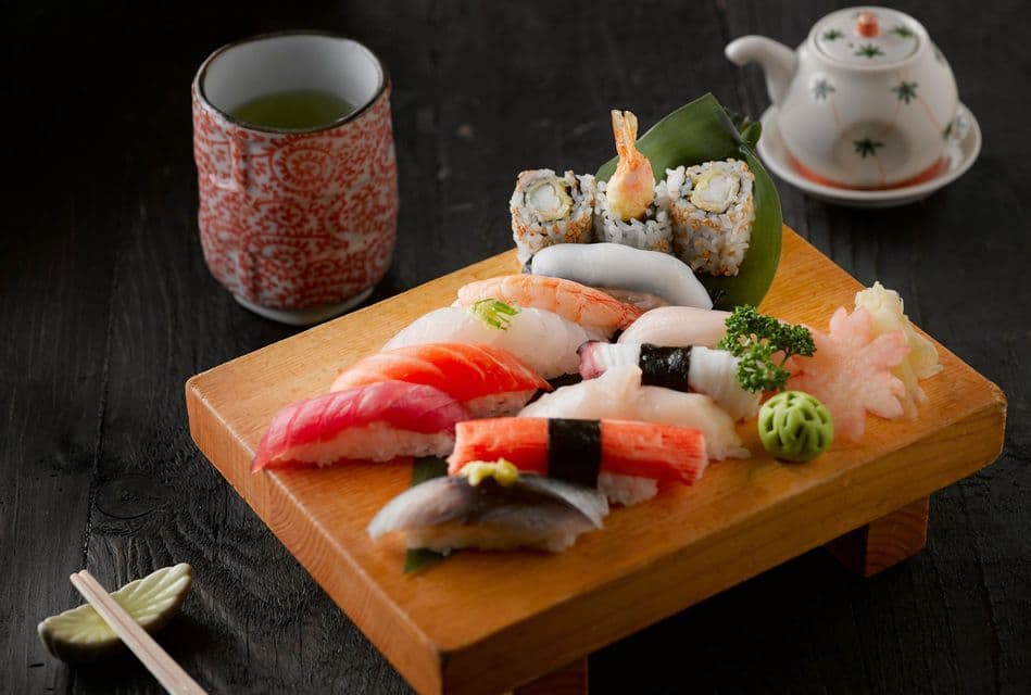 Una tabla de madera con sushi variado y rollos de maki se sirve con una taza de té verde y una tetera sobre una mesa oscura.