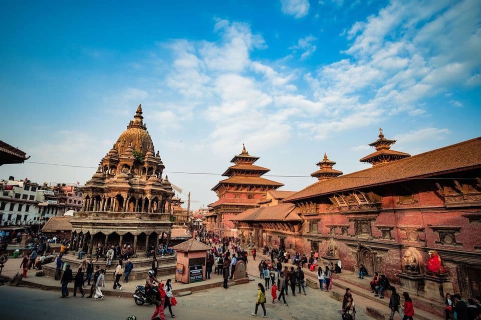 A crowd of people walks through a historic square filled with ornate temples and traditional buildings under a blue sky.