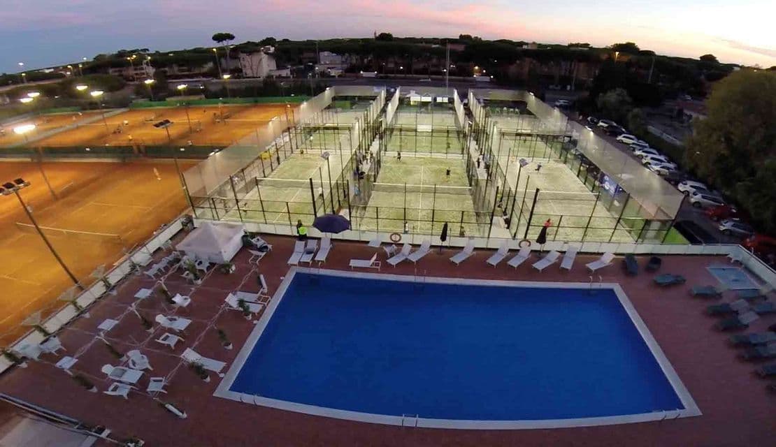 Una veduta aerea di un complesso sportivo al tramonto, con una piscina, diversi campi da padel illuminati e campi da tennis in terra battuta.
