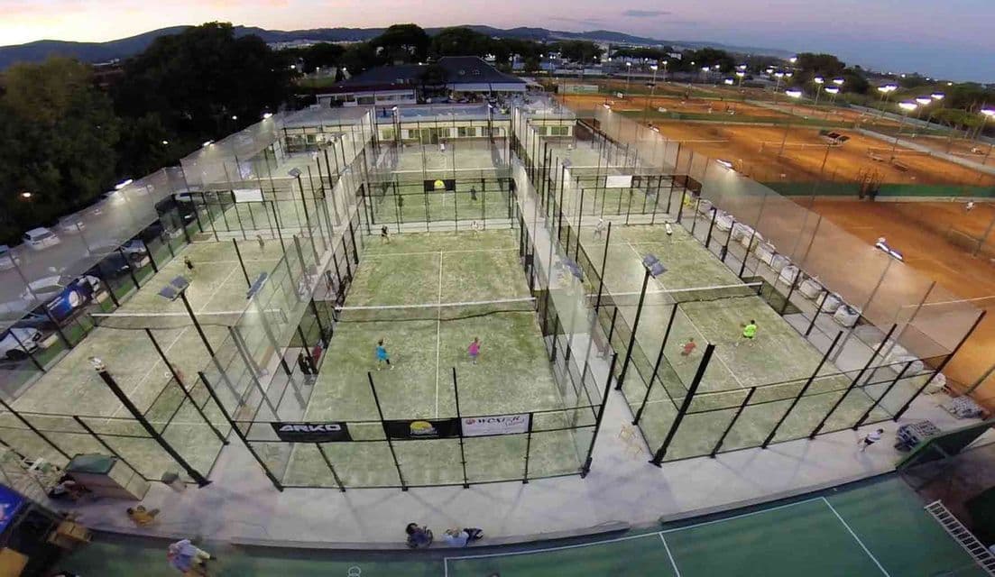 Una vista aerea di diversi campi da padel e tennis al tramonto, con persone che giocano sui campi da padel illuminati.
