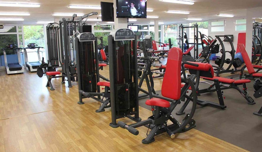 Una palestra vuota con varie macchine per pesi nere e rosse, tapis roulant e panche su un lucido pavimento in legno.