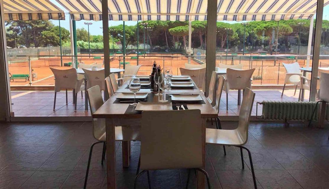Un tavolo da pranzo apparecchiato all'interno di un ristorante, con grandi finestre che si affacciano su campi da tennis in terra battuta vuoti.