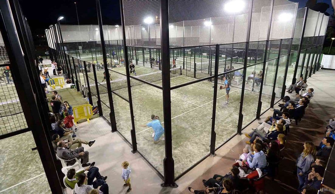 Vista dall'alto di persone che giocano a padel su campi illuminati di notte, con pubblico.