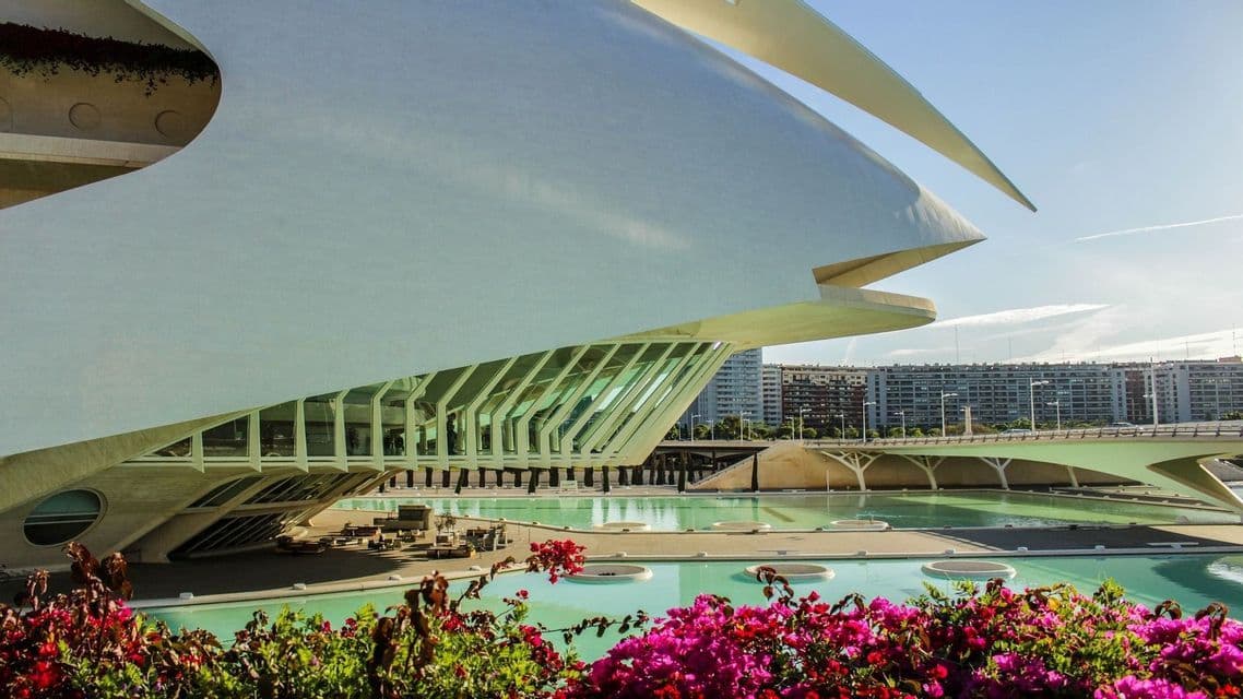 Un moderno edificio bianco con architettura curva accanto a una piscina turchese, con fiori rosa in primo piano.