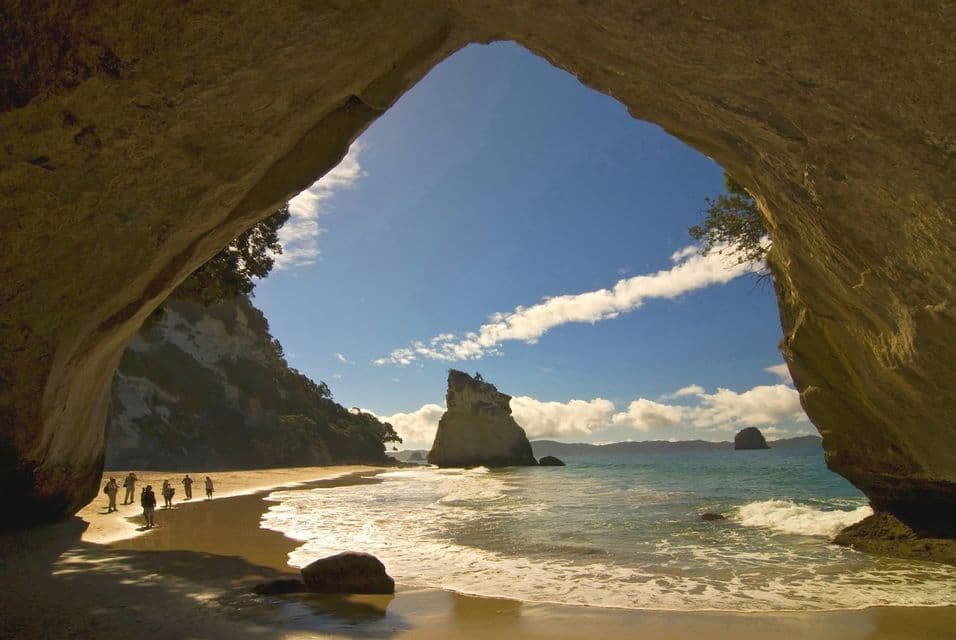 A WeRoad group trip walks on a wet sandy beach, framed by a large rock arch, with a sea stack in the sunlit ocean.