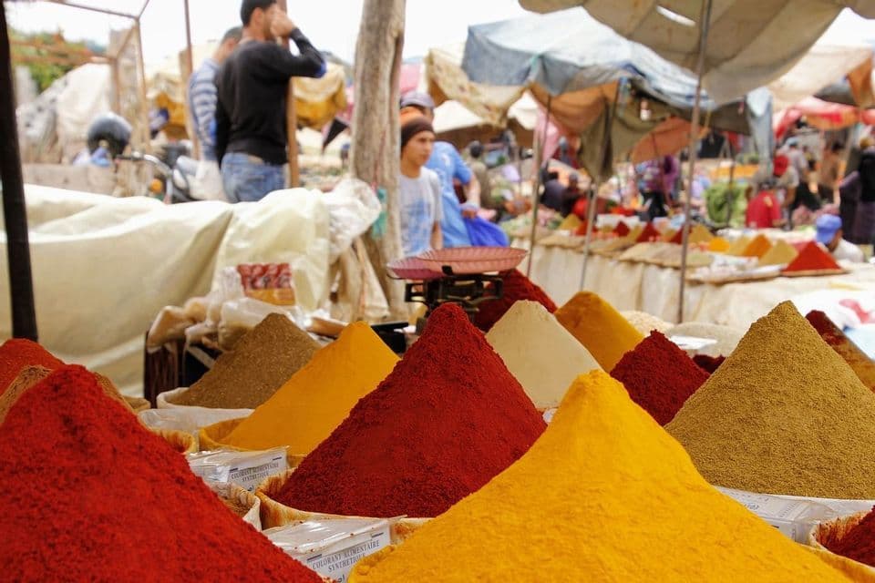 A close-up of various colorful spices arranged in conical piles at an outdoor market, with people blurred in the background.