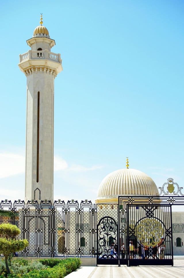 A white minaret and dome of a mosque stand behind an ornate metal gate under a bright blue sky.