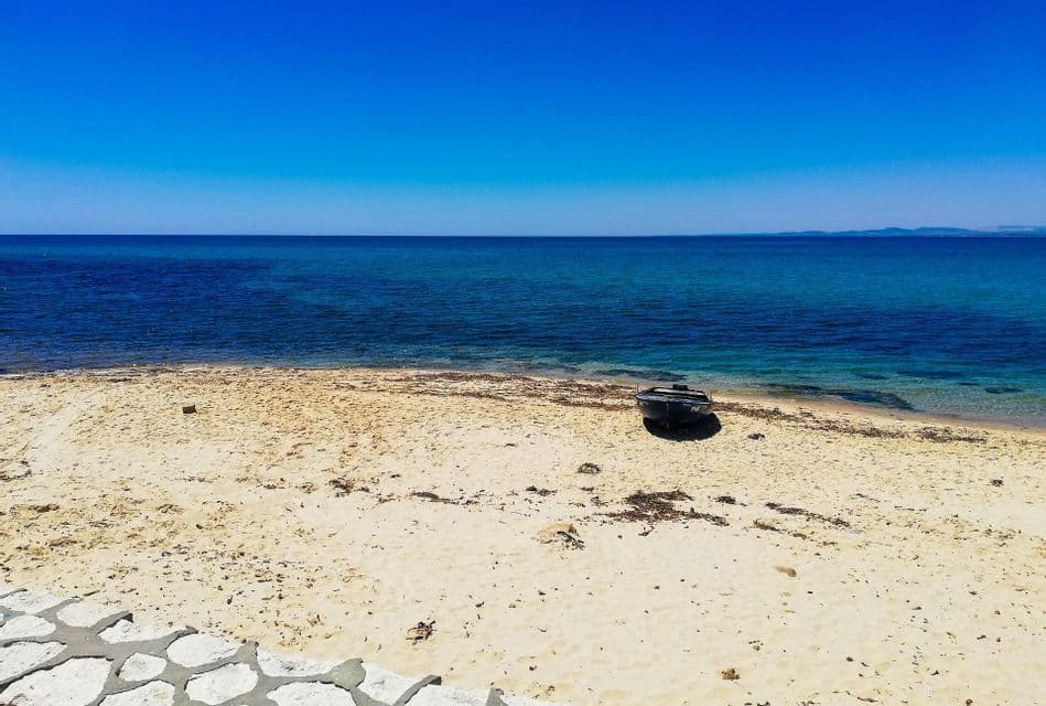 Un petit bateau sombre est échoué sur une plage de sable devant une mer calme et bleue sous un ciel sans nuages.