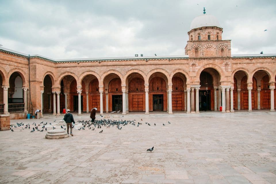 Two people walk in a large stone courtyard surrounded by an arched colonnade as pigeons gather on the ground.
