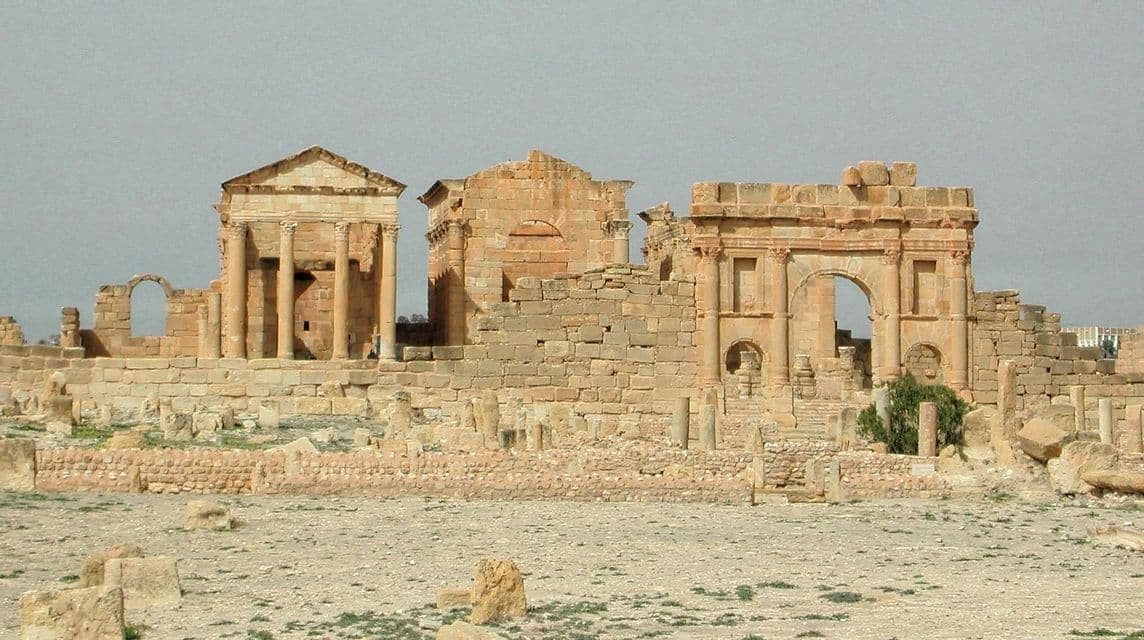 D'anciennes ruines de pierre, dont un temple et une arche, se dressent dans un vaste paysage aride sous un ciel voilé.