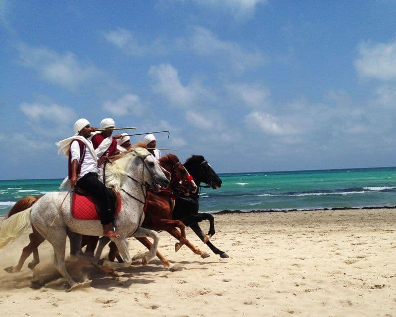 Four men in traditional attire galloping on horseback along a sandy beach next to the turquoise ocean under a blue sky.
