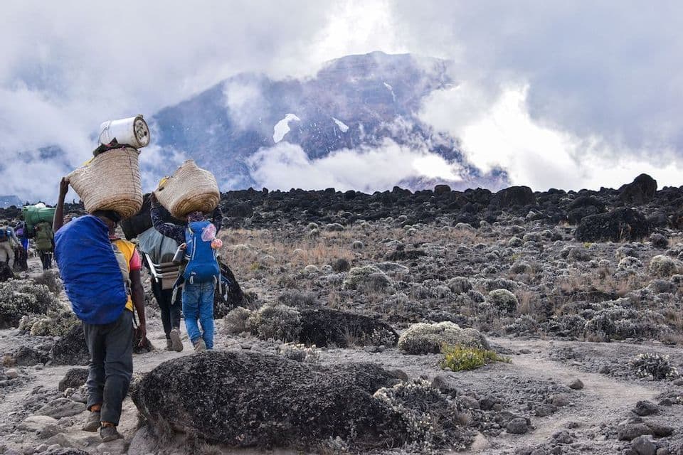 Un voyage de groupe WeRoad avec des porteurs transportant des paniers tressés marche sur un sentier rocheux vers un sommet de montagne couvert de nuages.