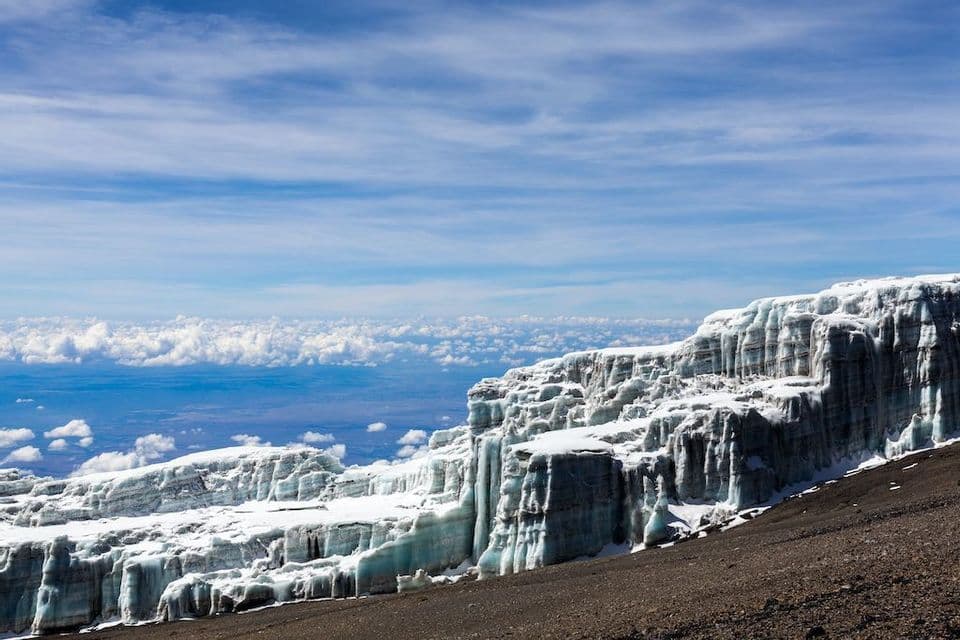 Un grand glacier avec des parois de glace verticales sur un versant rocheux de montagne, vu d'au-dessus d'une couche de nuages sous un ciel bleu.
