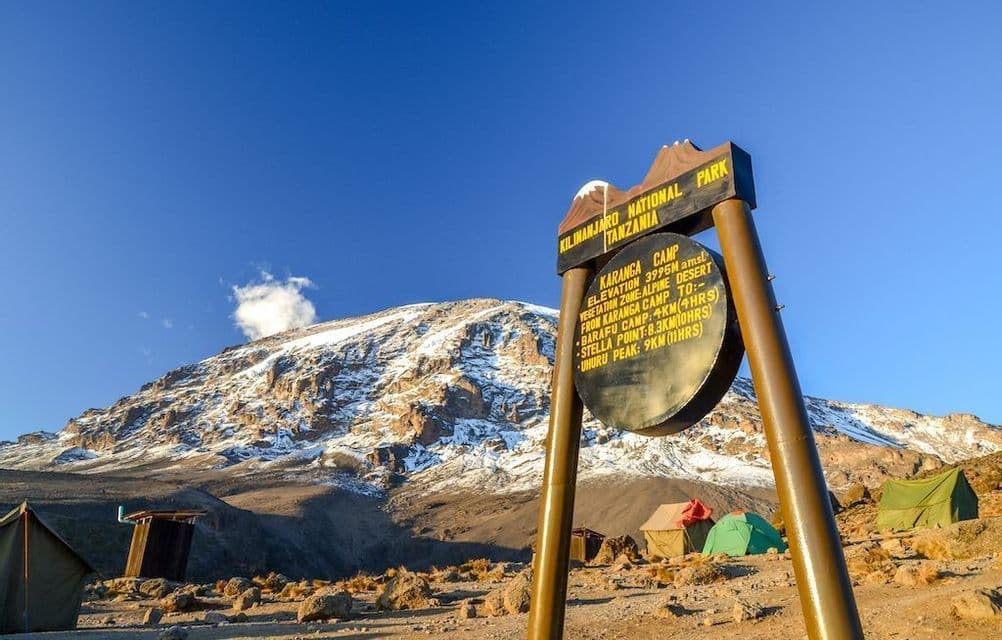 Un panneau pour le camp Karanga dans le Parc National du Kilimandjaro se dresse devant la montagne enneigée, avec des tentes plantées sur un terrain rocheux.