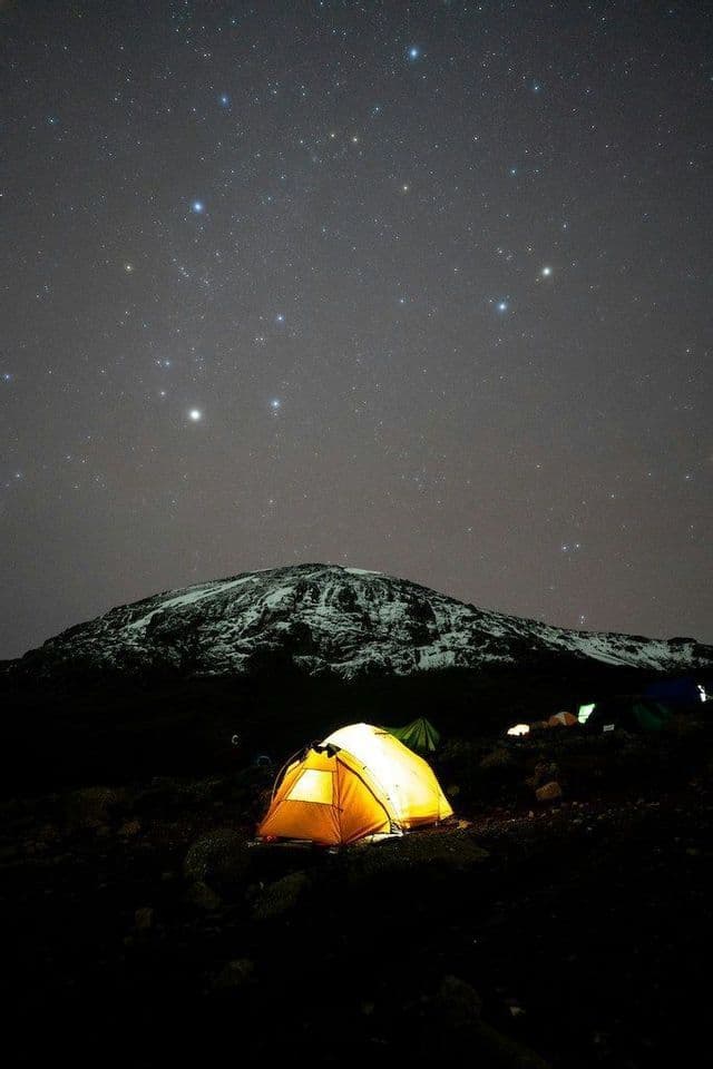 Une tente jaune illuminée sur un camping à flanc de montagne rocheuse sous un ciel nocturne étoilé, avec un sommet enneigé en arrière-plan.